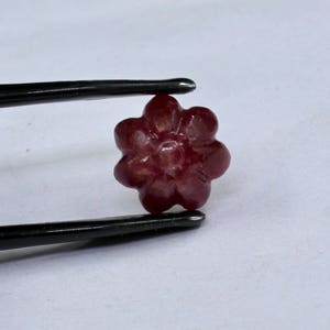 May include: A carved ruby gemstone in the shape of a flower. The flower has five petals and a center with a raised design. The gemstone is a deep red color.