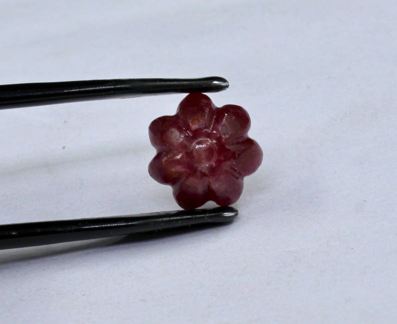 May include: A carved ruby gemstone in the shape of a flower. The flower has five petals and a center with a raised design. The gemstone is a deep red color.