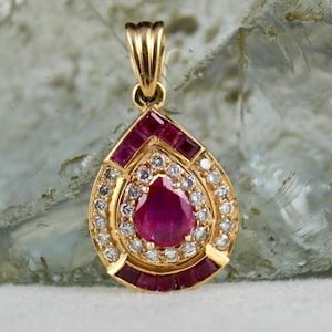 May include: A gold pendant featuring a pear-shaped ruby surrounded by a halo of smaller diamonds. The ruby is set in a gold teardrop-shaped frame with a border of smaller rubies.