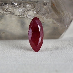 May include: A deep red, faceted gemstone with a marquise cut. The gemstone is set against a white background.