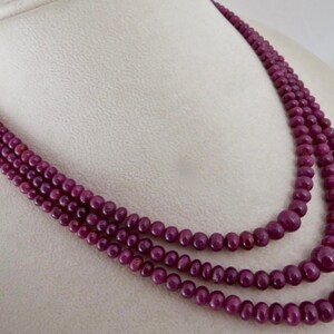 May include: A multi-strand necklace composed of deep red, round beads. The necklace is draped, showcasing the varying sizes of the beads and the rich color. The beads appear to be polished and evenly spaced, creating a luxurious look.