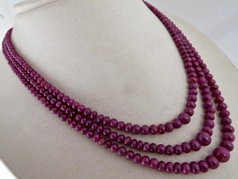 May include: A multi-strand necklace composed of deep red, round beads. The necklace is draped, showcasing the varying sizes of the beads and the rich color. The beads appear to be polished and evenly spaced, creating a luxurious look.