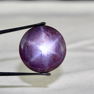 May include: A round, purple star ruby gemstone with a six-pointed star in the center. The gemstone is held by tweezers.