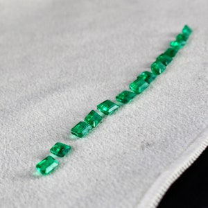 May include: A row of emerald green gemstones arranged in a line on a white surface. The gemstones are cut into rectangular shapes with beveled edges.