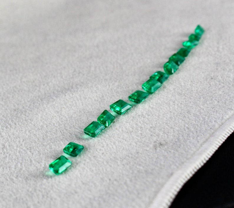 May include: A row of emerald green gemstones arranged in a line on a white surface. The gemstones are cut into rectangular shapes with beveled edges.