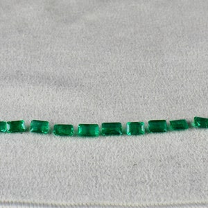 May include: A row of 15 emerald green rectangular gemstones of varying sizes arranged in a curved line on a white surface.