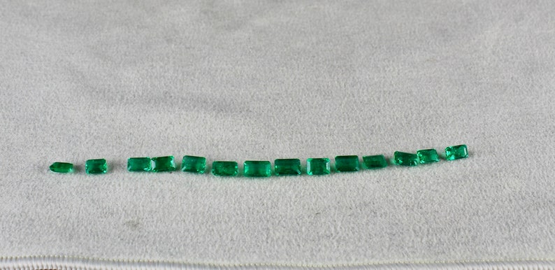 May include: A row of 15 emerald green rectangular gemstones of varying sizes arranged in a curved line on a white surface.