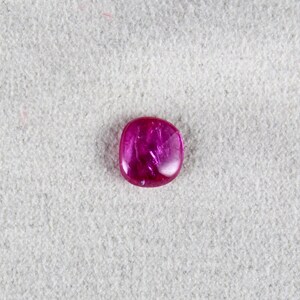 May include: A deep pink, faceted gemstone with a rounded, rectangular shape. The gemstone is set against a white, textured background.