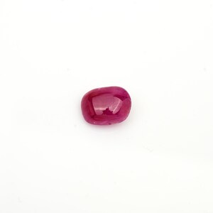 May include: A loose, oval-shaped ruby gemstone with a smooth, polished cabochon surface. The stone displays a deep, vibrant magenta-red color with translucent qualities and subtle internal inclusions visible through the polished dome.