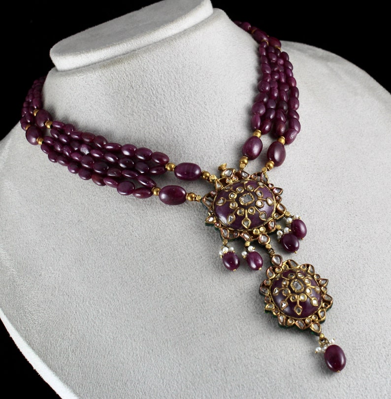 Pu&ograve; includere: Una collana a tre fili realizzata con perline viola scuro con un ciondolo in oro che presenta due disegni rotondi e decorati incastonati con pietre bianche.