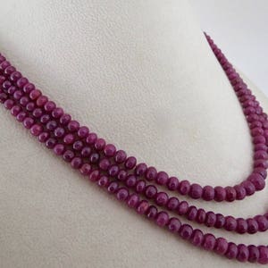 May include: A three-strand necklace composed of small, round, deep red beads. The necklace is draped over a white surface, showcasing the color and texture of the beads. The beads appear to be evenly sized and closely strung.
