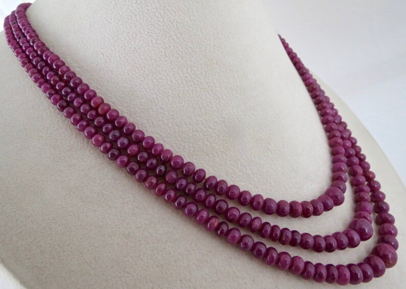 May include: A three-strand necklace composed of small, round, deep red beads. The necklace is draped over a white surface, showcasing the color and texture of the beads. The beads appear to be evenly sized and closely strung.