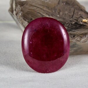 May include: A smooth, round, cabochon-cut gemstone with a deep red color. The gemstone is polished and has a glossy finish.