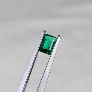 May include: A vibrant green emerald gemstone held in tweezers. The emerald is cut in a rectangular shape with facets.