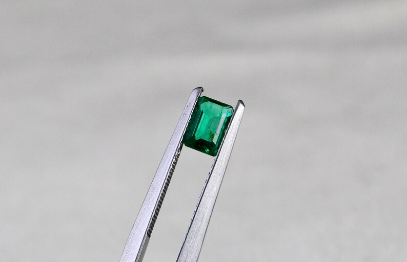 May include: A vibrant green emerald gemstone held in tweezers. The emerald is cut in a rectangular shape with facets.