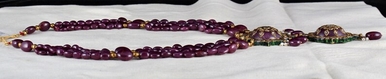 Pu&ograve; includere: Una collana multifilo realizzata con perline viola scuro con accenti dorati. La collana presenta due grandi ciondoli con disegni intricati e pietre preziose verdi.