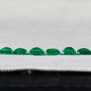 May include: A row of 17 emerald green gemstones of varying sizes, arranged in a line on a white fabric surface.