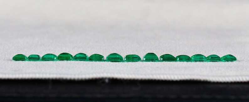 May include: A row of 17 emerald green gemstones of varying sizes, arranged in a line on a white fabric surface.