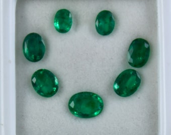 May include: Seven oval-shaped emerald green gemstones in a white box. The gemstones are arranged in a circle.