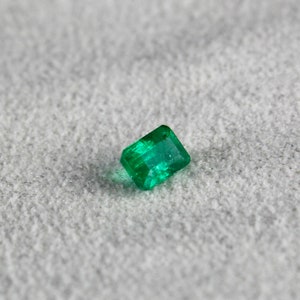 May include: A vibrant green emerald gemstone, cut into a rectangular shape, with facets that reflect light.