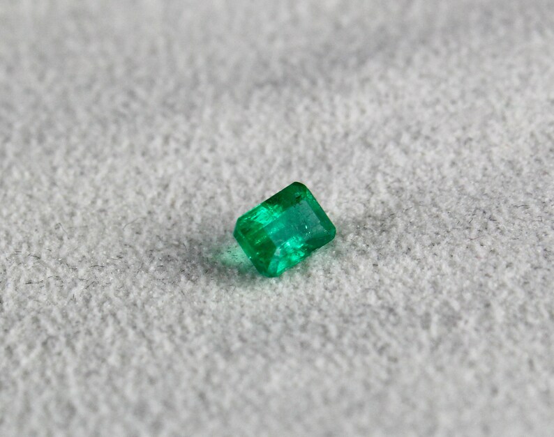 May include: A vibrant green emerald gemstone, cut into a rectangular shape, with facets that reflect light.