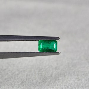 May include: A vibrant green emerald gemstone held in tweezers. The emerald is a rectangular cut with a smooth, polished surface.