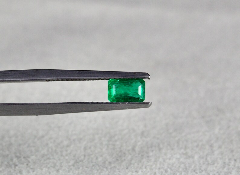 May include: A vibrant green emerald gemstone held in tweezers. The emerald is a rectangular cut with a smooth, polished surface.