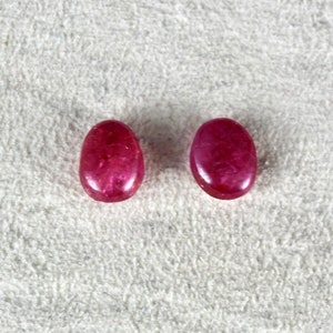 May include: Two oval-shaped, pink gemstones with a smooth, polished finish. The gemstones are set against a white, fuzzy background.