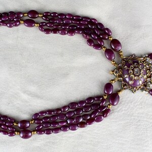 Pu&ograve; includere: Una collana multifilo realizzata con perline di pietre preziose viola con un ciondolo in oro che presenta una grande pietra preziosa rotonda viola circondata da pietre preziose bianche pi&ugrave; piccole. Il ciondolo &egrave; attaccato a un ciondolo rotondo in oro pi&ugrave; piccolo con pietre preziose bianche.