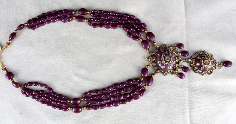 Pu&ograve; includere: Una collana multifilo realizzata con perline di pietre preziose viola con un ciondolo in oro che presenta una grande pietra preziosa rotonda viola circondata da pietre preziose bianche pi&ugrave; piccole. Il ciondolo &egrave; attaccato a un ciondolo rotondo in oro pi&ugrave; piccolo con pietre preziose bianche.