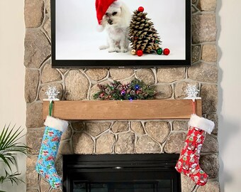 Op de afbeelding: Een schouw die versierd is voor kerstmis met een witte hond die een kerstmanmuts draagt ​​op het tv-scherm boven de schouw. Er hangen twee kousen aan de schouw, een blauwe en een rode.