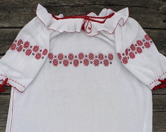 May include: White embroidered blouse with red floral pattern and a ruffled neckline.