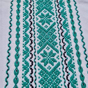 Könnte beinhalten: Nahaufnahme eines weißen Textils mit aufwendiger Stickerei. Das Design zeigt ein sich wiederholendes Muster aus türkisfarbenen und schwarzen geometrischen Formen und floralen Motiven. Die Stickerei ist detailliert und erzeugt eine strukturierte Oberfläche.