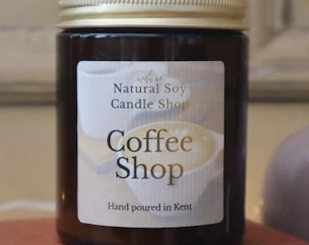 May include: A dark amber glass candle jar with a gold metal lid. The label features a graphic of a coffee cup and the text Natural Soy Candle Shop, Coffee Shop, and Hand poured in Kent. The candle is contained within a cylindrical glass vessel.