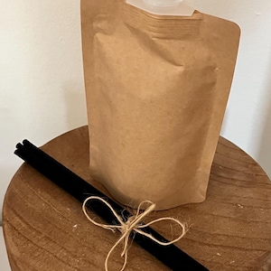 Könnte beinhalten: Ein brauner Kraftpapierbeutel mit weißem Kunststoffverschluss, der auf einer Holzoberfläche liegt. Schwarze Diffusorstäbchen liegen neben dem Beutel und sind mit Bindfaden gebunden. Der Beutel ist wahrscheinlich für flüssige Produkte.
