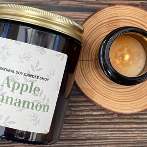 May include: A brown glass candle jar with a gold lid and a white label that reads "NATURAL SOY CANDLE SHOP Apple Cinnamon". The candle is lit and the wax is melting.