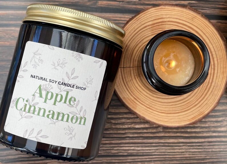 May include: A brown glass candle jar with a gold lid and a white label that reads "NATURAL SOY CANDLE SHOP Apple Cinnamon". The candle is lit and the wax is melting.