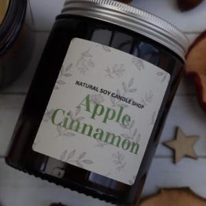 May include: A brown glass candle jar with a white label that reads "Natural Soy Candle Shop" and "Apple Cinnamon". The candle is surrounded by cinnamon sticks, dried apple slices, and wooden stars.