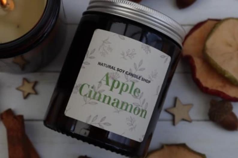 May include: A brown glass candle jar with a white label that reads "Natural Soy Candle Shop" and "Apple Cinnamon". The candle is surrounded by cinnamon sticks, dried apple slices, and wooden stars.