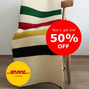 May include: A cream-colored blanket with horizontal stripes in green, red, yellow, and black is draped over a wooden chair. The blanket has a black stitched border. The image also includes a red circle with the text "Buy 1, get 2nd 50% OFF".