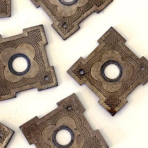May include: A collection of square metal plates with a weathered bronze finish. Each plate features a central circular opening surrounded by a four-lobed clover shape, with textured stippled detailing on the surface and small screw holes.