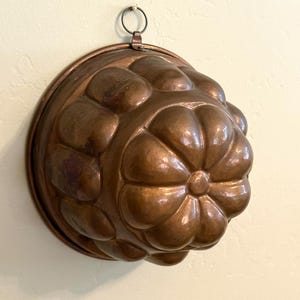 May include: A round copper mold featuring a raised, multi-lobed floral or petal design across its surface. The metallic object has a warm, reddish-brown finish and includes a small, circular metal loop attached to the top edge for hanging.