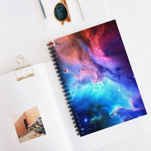 Op de afbeelding: Spiraalgebonden notitieboek met een levendig kosmisch nevelontwerp in blauwe, paarse en rode tinten. Het notitieboek is open en toont witte pagina's en een zwarte spiraalbinding. Een gouden paperclip en zonnebril zijn ook zichtbaar.