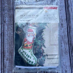 May include: A cross-stitch kit for a Christmas stocking. The stocking features a Santa Claus design with the words "Merry Christmas." The kit is packaged in a clear plastic bag with the brand name "Bernat" visible.