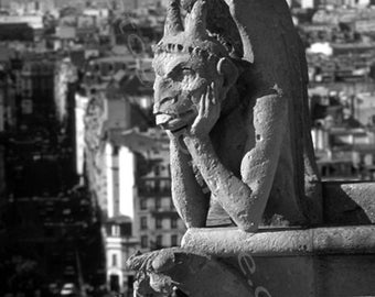 May include: A black and white photo of a gargoyle sculpture perched atop a building. The gargoyle is carved in a seated position with its head resting on its hand, looking out over the city.
