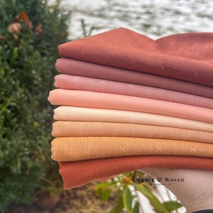 Peut inclure: Une pile de carrés de tissu dans différentes nuances de rouge, de rose et d'orange. Le tissu présente un motif subtil de petites croix. L'image est prise à l'extérieur avec un arrière-plan vert flou.