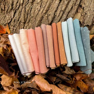 May include: A collection of folded fabric bundles in various pastel colors, including white, peach, and teal, arranged on a bed of autumn leaves. Each fabric piece features a subtle cross-stitch pattern. The text "Weave & Woven" is visible.