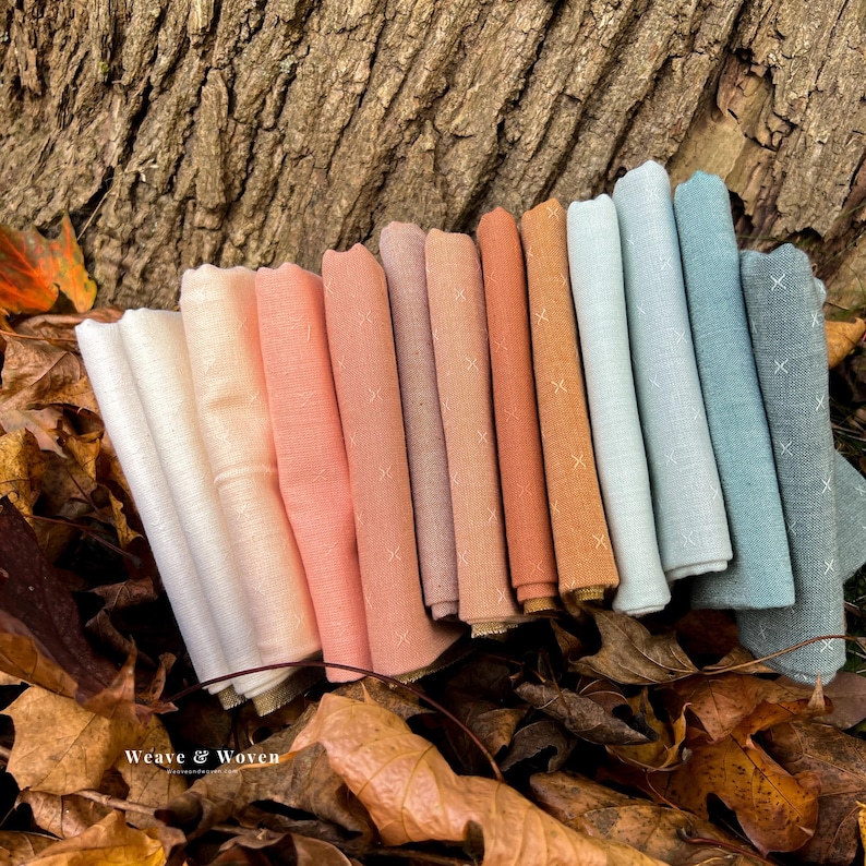 May include: A collection of folded fabric bundles in various pastel colors, including white, peach, and teal, arranged on a bed of autumn leaves. Each fabric piece features a subtle cross-stitch pattern. The text "Weave & Woven" is visible.