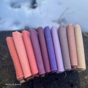 Peut inclure: Une collection d'échantillons de tissu roulés dans un dégradé de couleurs, allant du corail au beige. Les tissus sont disposés en éventail, mettant en valeur leurs différentes teintes et textures. Le texte "Weave & Woven" est visible en bas à gauche.