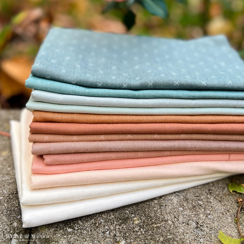 May include: A stack of folded fabric in various colors, including shades of blue, green, peach, and cream. The fabric has a subtle cross-stitch pattern. The image is well-lit, showcasing the textures and colors of the textiles. The bottom left corner shows the text "Weave & Woven".